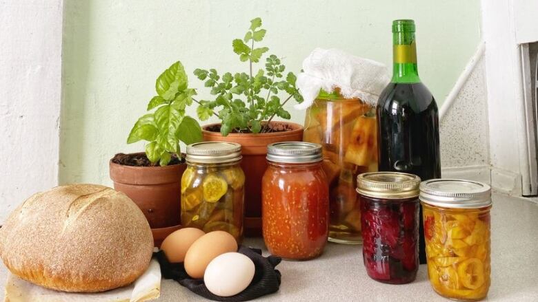 A loaf of bread, eggs, herbs in pots, and jars of various foods sit on a counter.