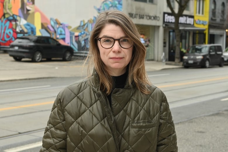 Close-up of woman wearing olive green quilted coat standing on the sidewalk. An empty city street and a row of businesses are behind her. 