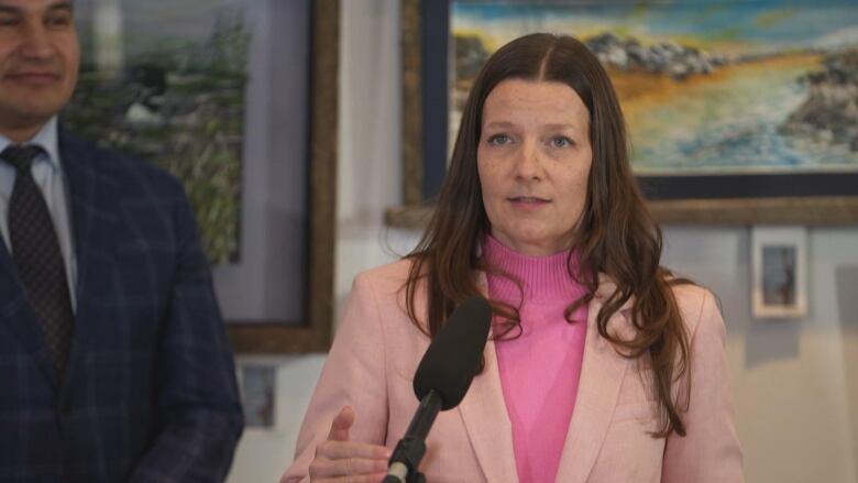 A woman with shoulder-length brown hair in a pink sweater and pink blazer speaks to media in front of a lectern.