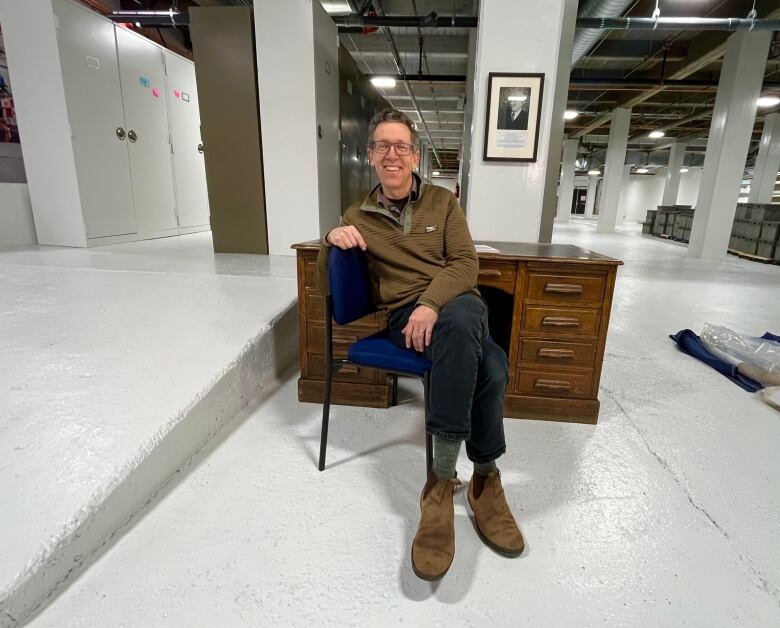 A smiling man sits in a barren industrial space at an antique oak desk. 