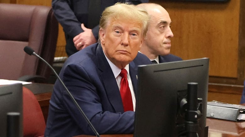 An older, clean-shaven man in a suit and tie looks toward the camera while sitting at a table inside a court beside another man.