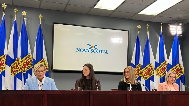 Four women sit in front of microphones at a news conference.