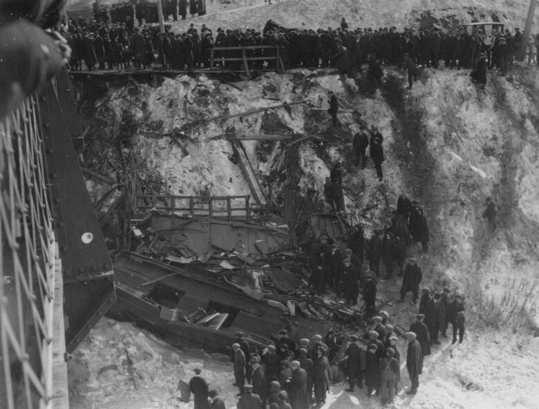 An old black and white photo shows a large crowd gawking at the twisted remains of a streetcar lying in a riverbed below a bridge.