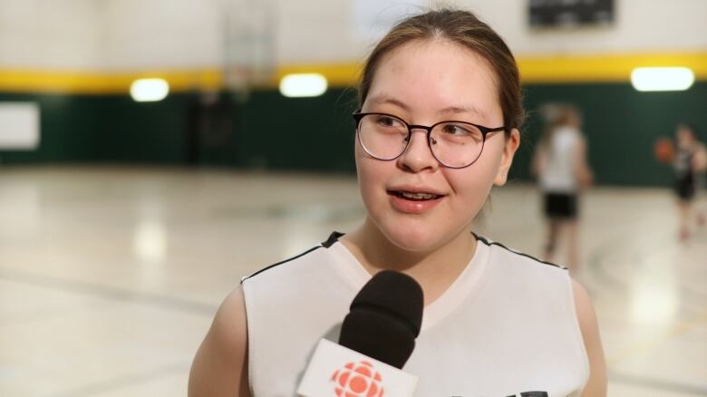 A girl with glasses in a white basketball uniform smiles while talking into a microphone