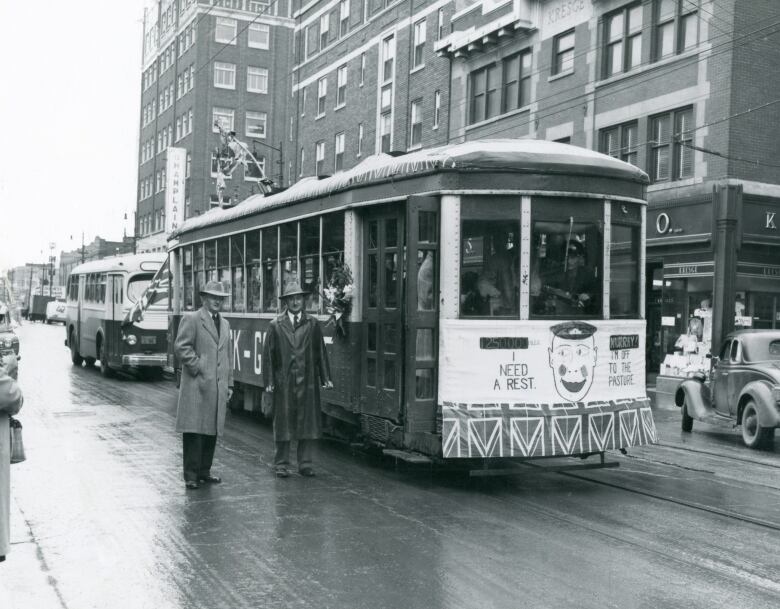 An old black and white photo shows two men in trenchcoats and hats standing in front of a streetcar in Regina.