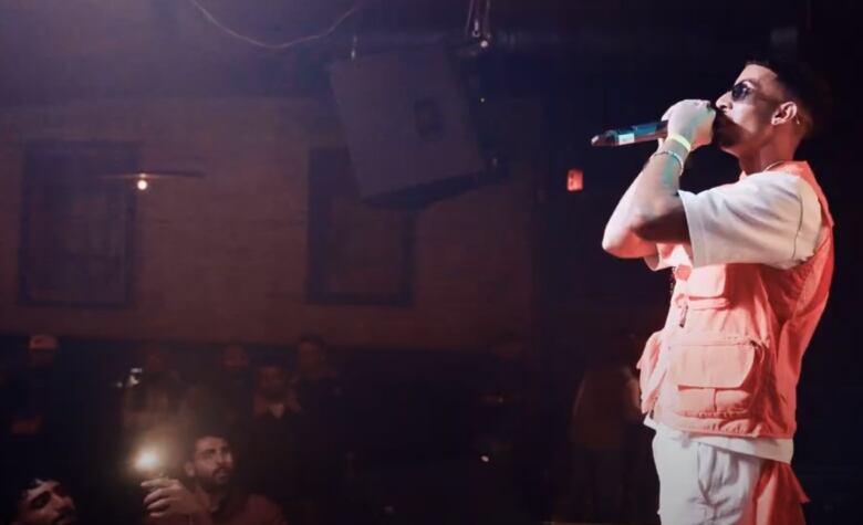 A man with black hair, wearing an orange vest over a white T-shirt, sings onstage into a microphone in front of a crowd.