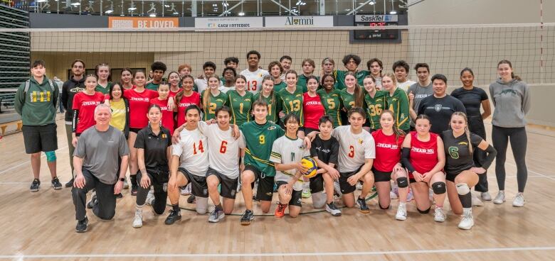 A large collection of young volleyball players pose with coaches in from of a volleyball net.