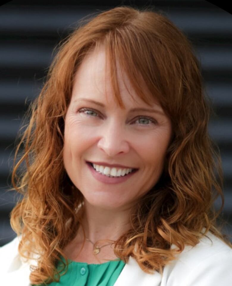 A woman with curly auburn hair in an emerald blouse with a white jacket smiles at the camera.