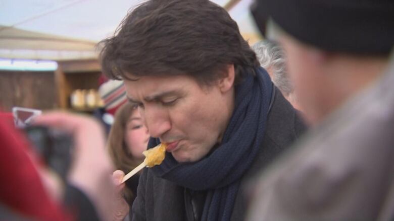 A man with short hair, wearing a black scarf and jacket, is shown eating some taffy off of a stick.