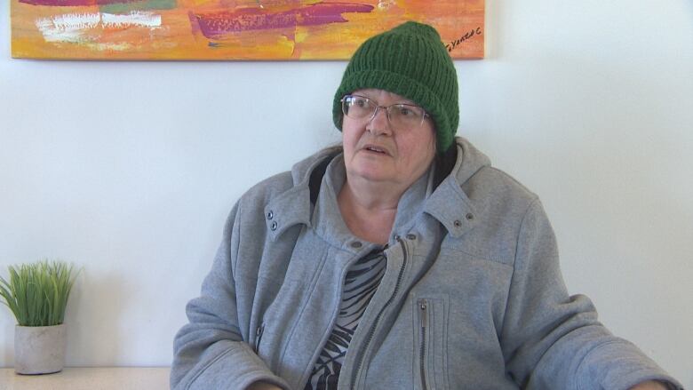 A woman wearing a green toque sits on a chair in front of a wall.