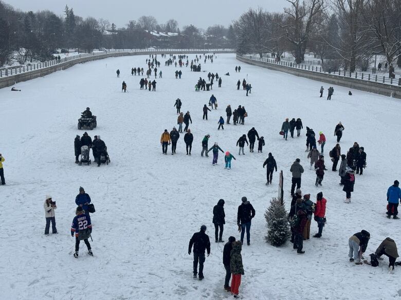 People skate and walk on the Rideau Canal Skateway. The ice is covered in snow and its a cloudy day.