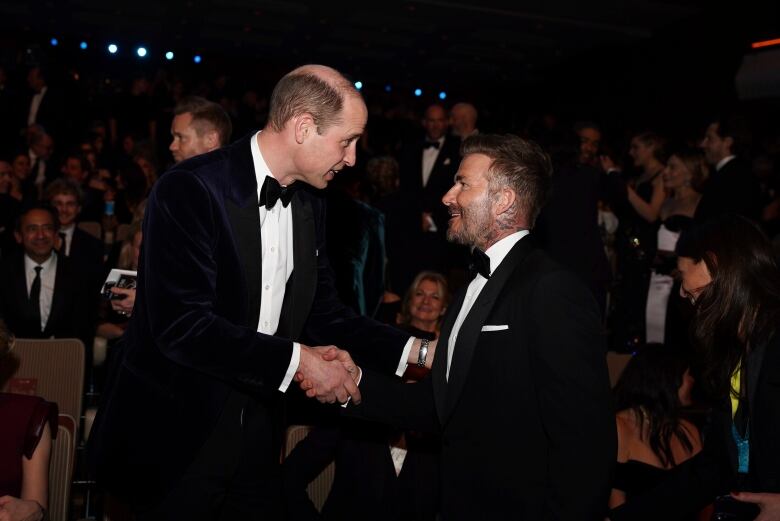 Two people in tuxedos shake hands as people sit in an audience behind then.