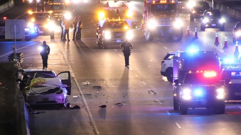 Emergency cars flash their lights around a pair of crashed vehicles located on the shoulder of a highway.