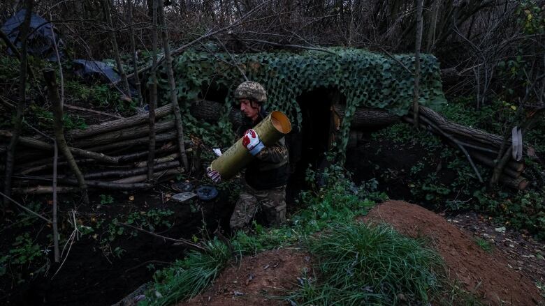 A soldier holds a large shell with both arms near camouflage.