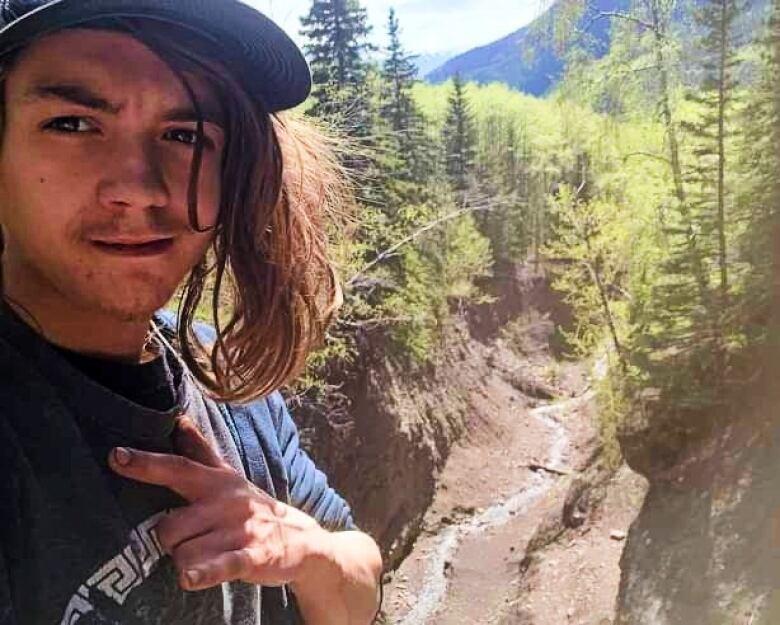 A young man with long hair and a ball cap looks into the camera, with a mountain scene behind him.