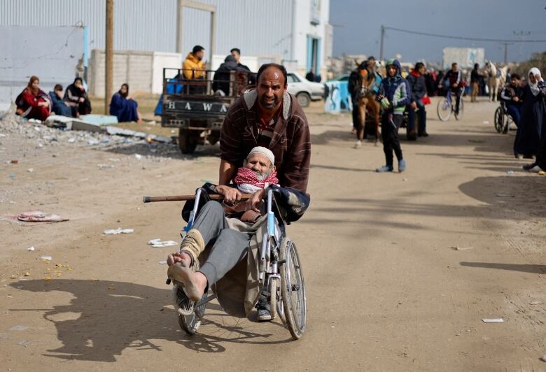 A man pushes an older man in a wheelchair down a dirt road. The older man is carrying a cane. His bare feet are dirty and one leg is bandaged. Behind them, a crowd of people moves in the same direction by foot, bicycle, cart or vehicle. Some people sit on the side of the road.