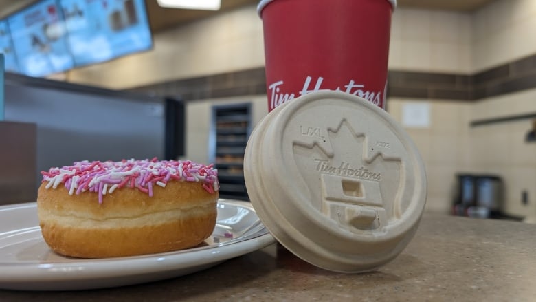 A paper coffee cup lid is seen in front of a red tim hortons cup. On the left is a sprinkle donut on a plate.