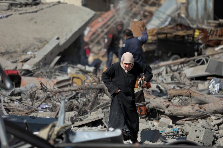 A woman wearing a head covering and long coat walks over concrete rubble and debris.