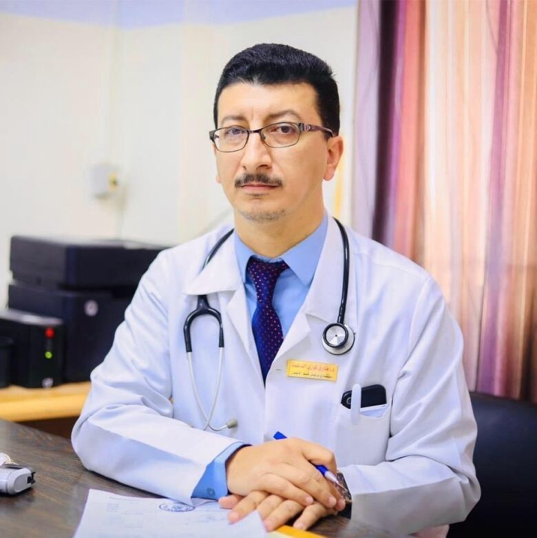 A mustachioed man in a white lab coat with a stethoscope around his neck sits at a desk with his hands folded in front of him and a serious expression on his face. 