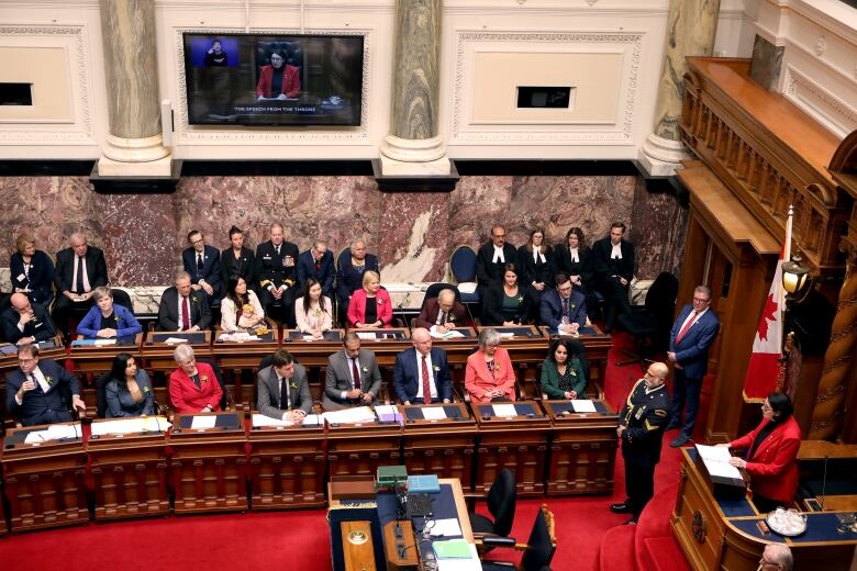 Rows of people sit and listen to a woman deliver a speech in a legislature building.