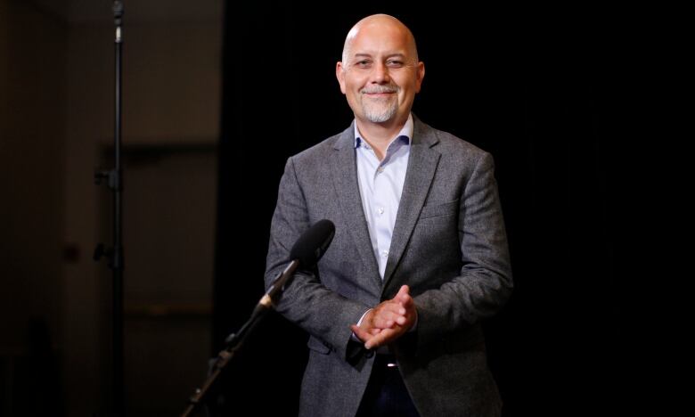 A bald Indigenous man with a white goatee wearing a grey jacket smiles while standing in front of a mic.