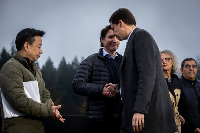 An East Asian man stands at the side as two tall white men shake hands, with a white woman and Indigenous man in the background.