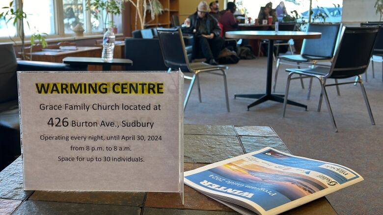 A sign on a table in a library reads 'Warming Centre' with patrons in the background