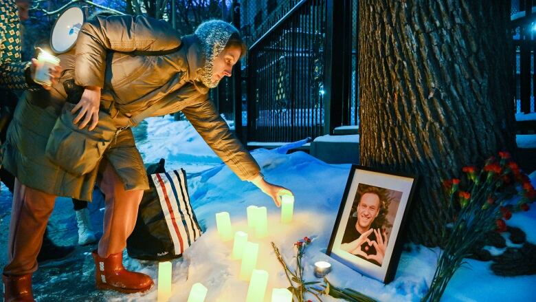 A woman in a winter coat lays a candle in front of a framed photo of a man. The photo is leaning up against a tree. Flowers lay beside the photo.