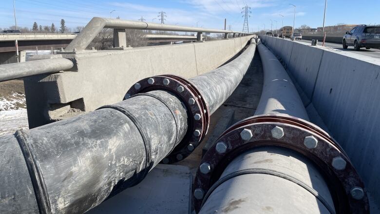 Long pipes run along the side of a bridge away from the camera. Cars can be seen driving over the bridge.