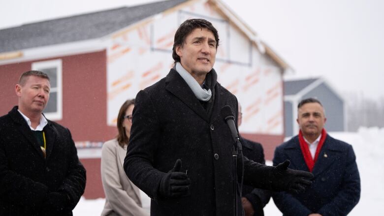 The prime minister stands in a coat outside with snow and officials in the background.