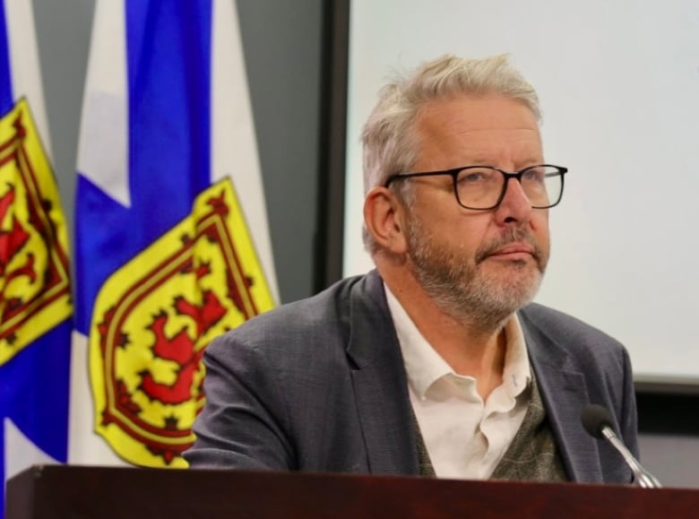 A man with white hair and wearing a grey suit sits in front of a Nova Scotia flag.