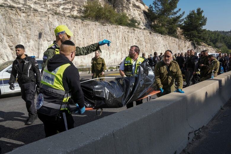 Two men in bright yellow vests stand at either end of a stretcher with a black body bag strapped to it, while other men in green uniforms stand nearby along a road.