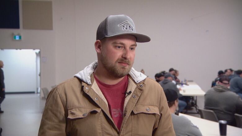 A man in a brown coat, red t-shirt and grey baseball hat stands in front of tables.