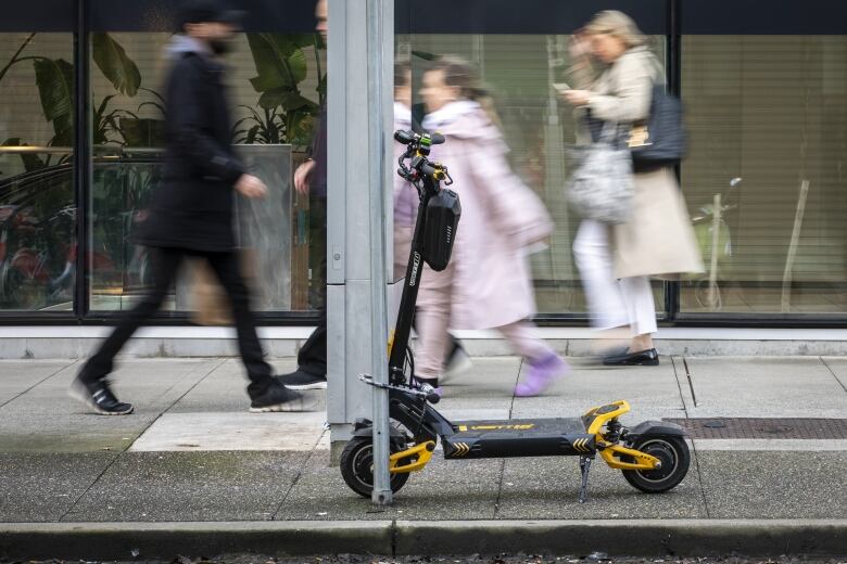 An e-scooter is seen parked and locked against a pole amid a busy side, with blurred images of people walking in the background. 