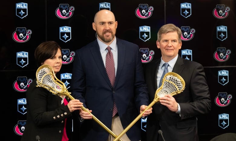 Three sports executives pose with lacrosse sticks at a news conference.