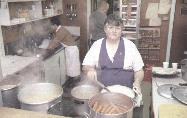 A vintage photo of a matronly woman in a purple apron cooking a large vat of hot dogs. 