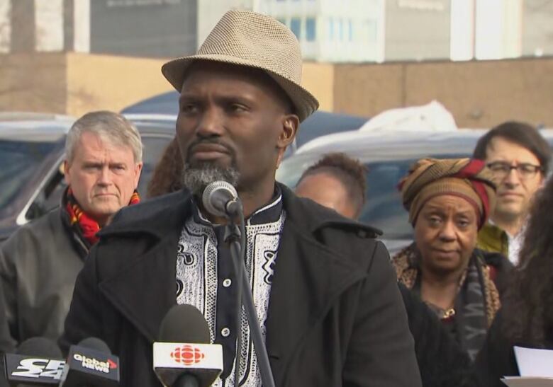 A middle-aged Black man in a fedora stands in front of several microphones outside on a cold morning. About half a dozen people can be seen standing behind him.