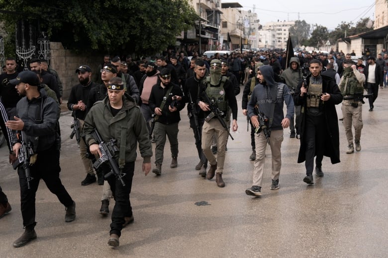 Several men march in an urban setting, several are carrying long guns on straps across their bodies.