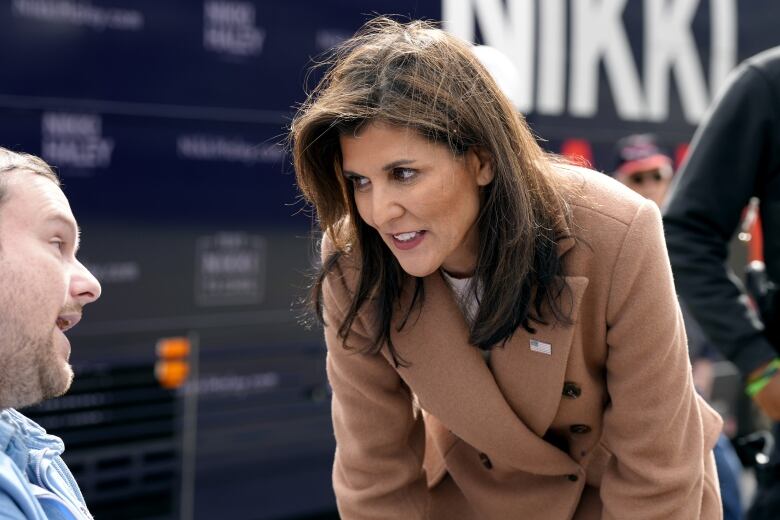 A woman with dark hair wearing a tan coat leans over to speak with a supporter. 