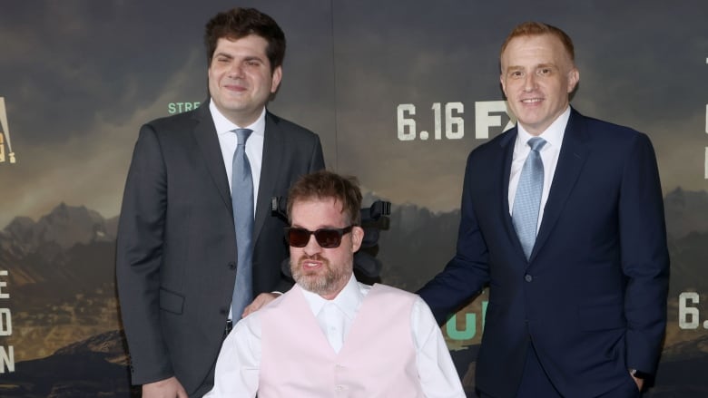 A man seated in a wheelchair is shown between men who are standing and wearing suits and ties.