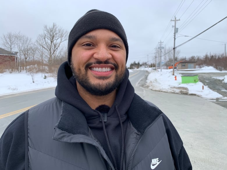 A Black man with a short beard wearing a black tuque and hoodie stands outdoors in the winter time. 