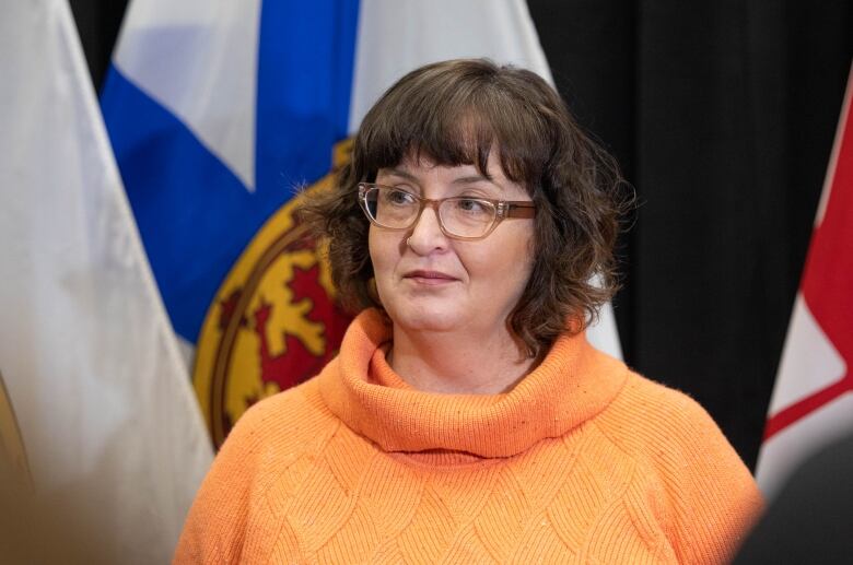 A personj with glasses and dark hair stands in front of flags.