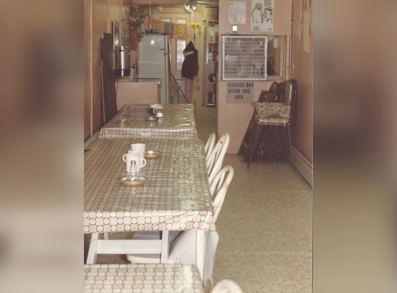 A vintage 1980s photo of a basic soup kitchen dining room with a small kitchen and a few tables.