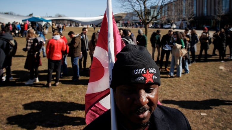 A man with a flag stands in front of people gathered at a rally.