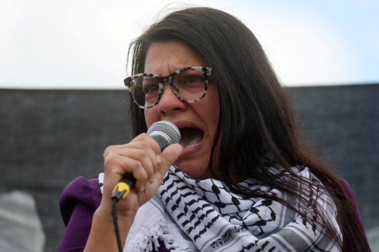 A woman with long brown hair, leopard-print glasses and a keffiyeh shouts into a microphone