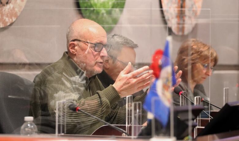 A man wearing glasses and a green sweater gesturing behind a desk surrounded by Plexiglas.