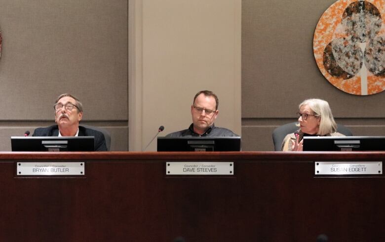 Three people behind a dark brown desk with a man on the left wearing glasses looking at the ceiling.