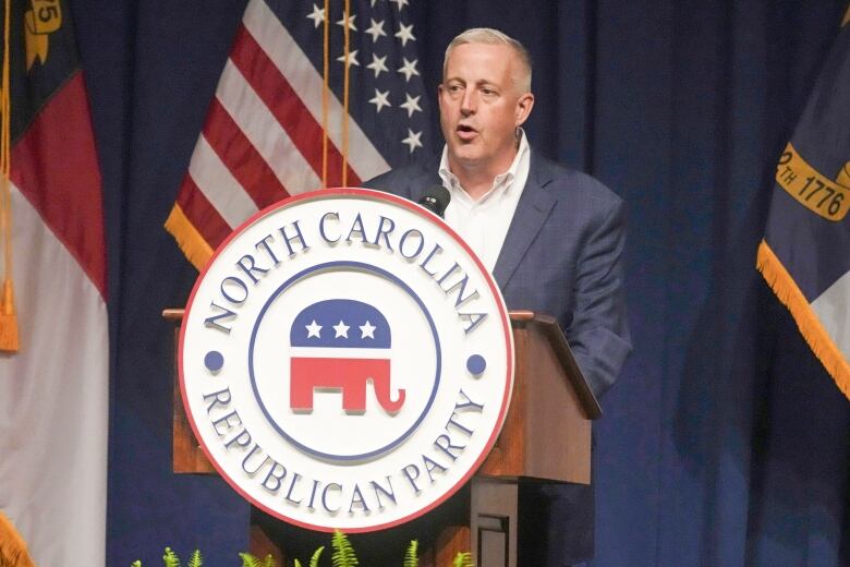 A clean-shaven man in a suit jacket and collared shirt is shown speaking at a podium that says 'NOrth Carolina Republican Party.'