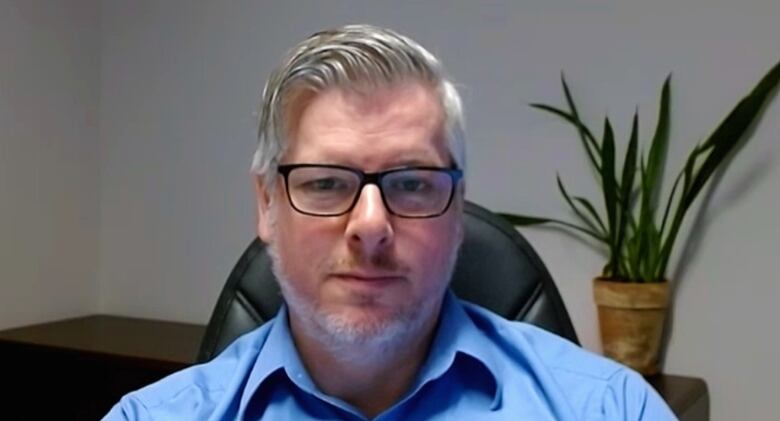 A man in glasses and a blue button-up shirt sits in an office with a plant behind him. 