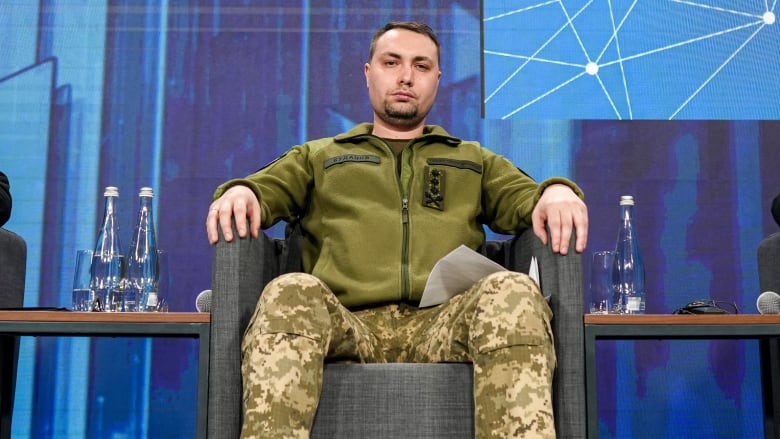 A man in military fatigues sits in a chair on a stage.